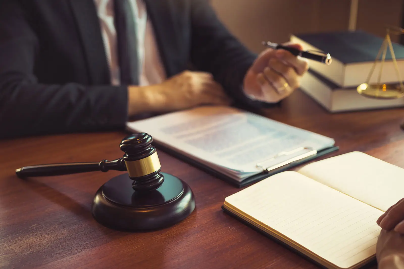 a lawyer reaches a pen to a client to sign a legal document. A gavel with sound block, books, and a justice scale is on top of the wooden table.