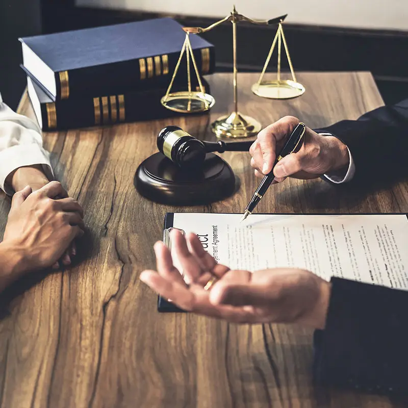 A lawyer explains the content of a legal document to a client. A gavel, scale of justice and two books are on top of the wooden table.