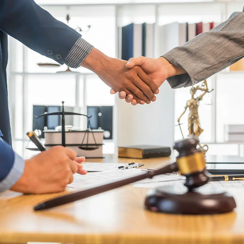 laywer and client shaking hands. A justice scale and gavel with sound block is on top of the wooden table.