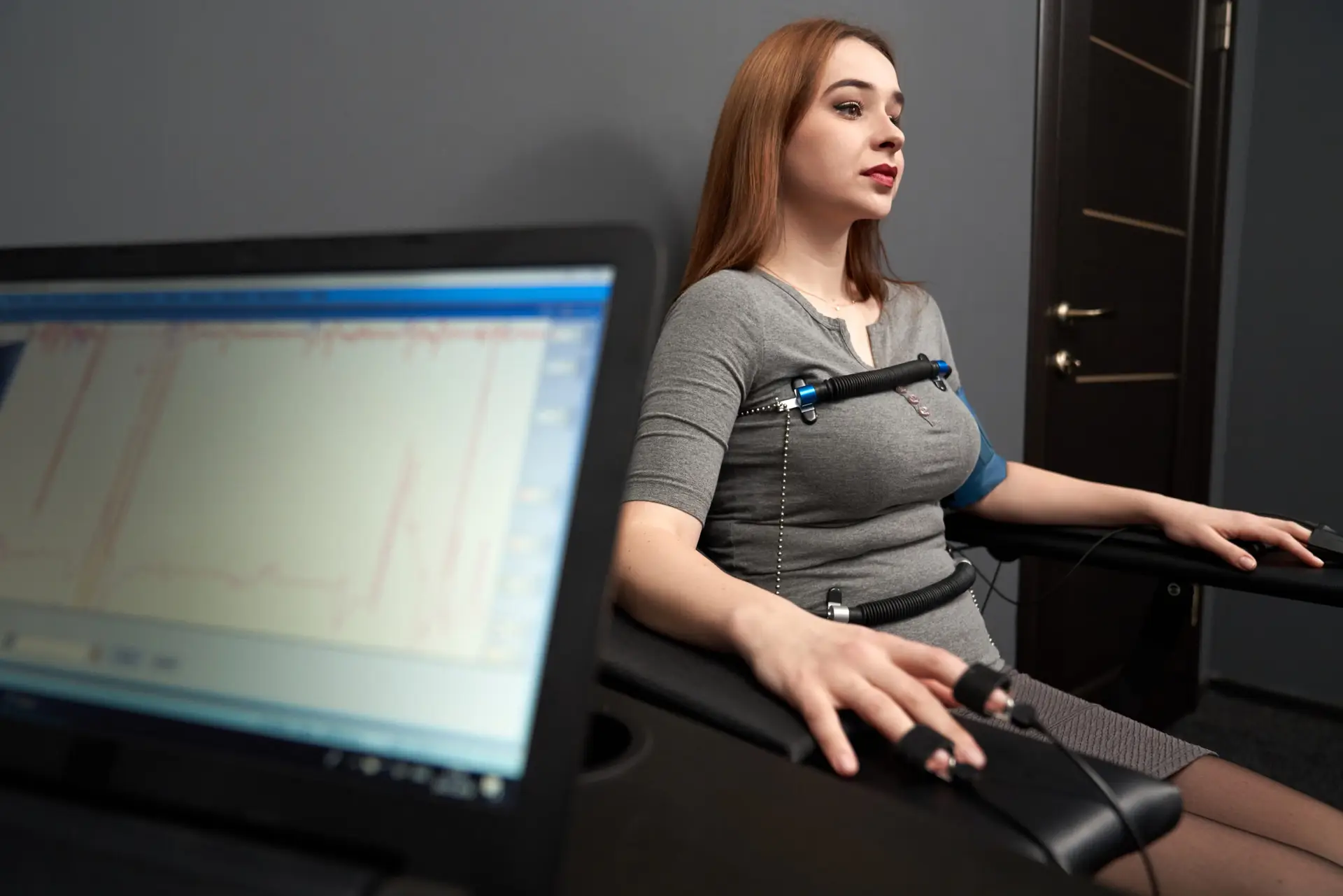 Young beautiful woman sitting with attached sensors on fingers, hands and body, testing with computer polygraph, looking away. Graph and polygraph on screen of lie detector.