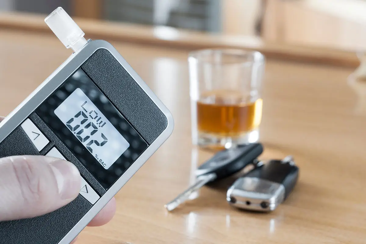 A man is holding a breathalyzer test with a low reading. Car keys and a glass filled with liquor is on top of a wooden table.